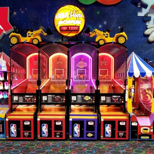 Three basketball arcade games with NBA branding, featuring yellow toy trucks on top, set in a colorful game room.