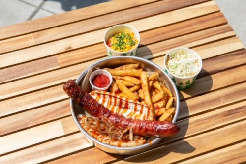 A bowl of grilled chicken, sausage, fries, and two cups of sides on a wooden table.