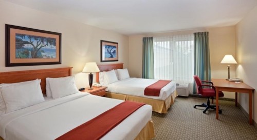 A cozy hotel room with two beds, a desk, and a window with curtains, featuring warm lighting and artwork on the walls.