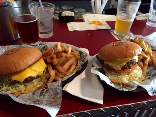 Two cheeseburgers with lettuce and tomato, served with a side of golden fries, on a red table with drinks in the background.