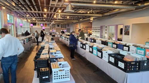 A spacious record store with customers browsing through rows of vinyl records in bins and on tables.