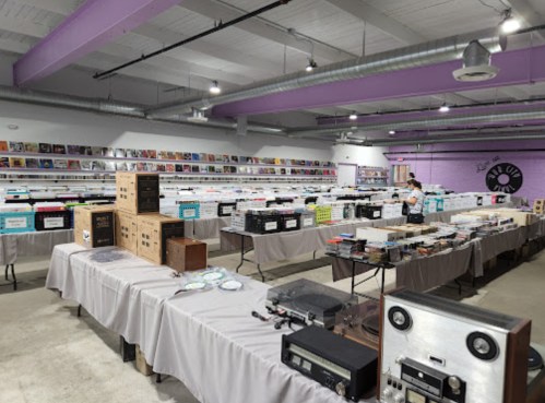 A spacious record store with tables displaying vinyl records, audio equipment, and boxes, under bright lighting.