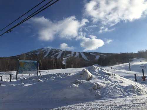 Snow-covered mountain with ski trails, blue sky, and clouds; a map sign and snowbank in the foreground.