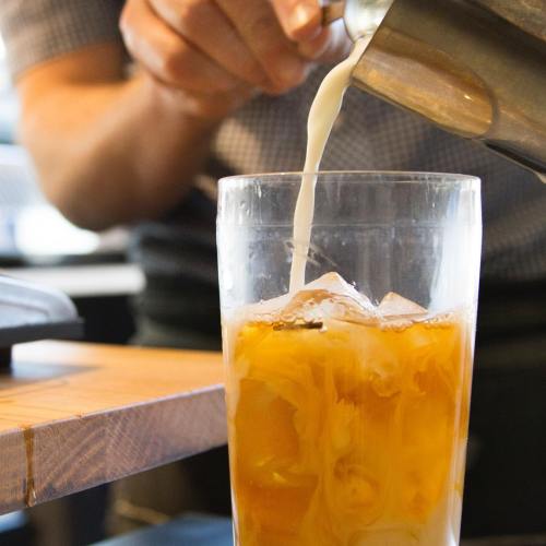 A person pours milk into a glass of iced tea, creating a swirl effect with ice visible in the drink.