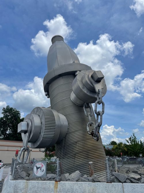 A large, metallic fire hydrant sculpture stands against a blue sky with fluffy clouds, surrounded by a chain-link fence.