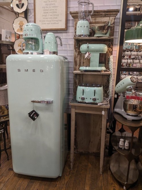 A vintage-style mint green SMEG refrigerator and kitchen appliances displayed on a wooden shelf in a cozy store.