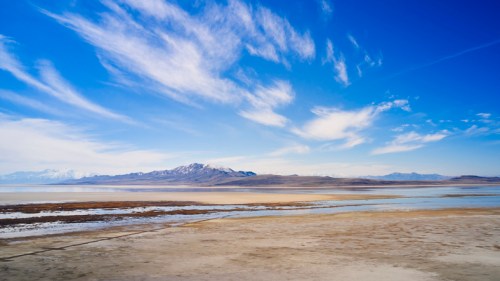 A vast landscape featuring a flat, dry area with mountains in the background under a bright blue sky with wispy clouds.