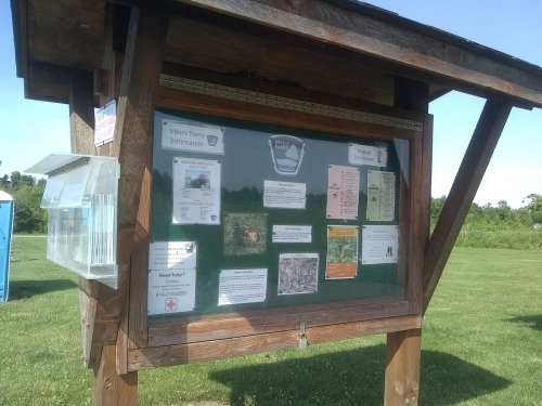 A wooden bulletin board displaying various informational flyers and a clear box for inquiries, set in a grassy area.