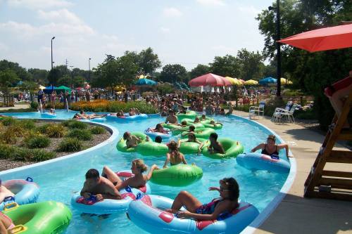 A busy water park with people floating on inner tubes in a lazy river, surrounded by colorful umbrellas and greenery.