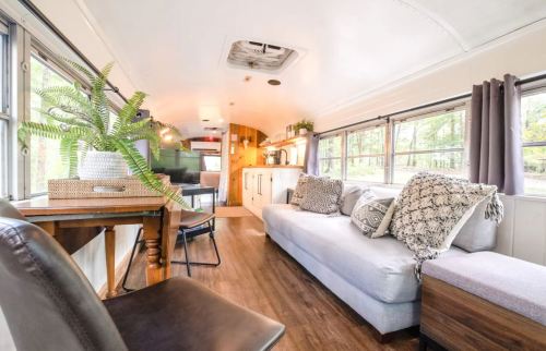 Cozy interior of a converted bus, featuring a sofa, dining table, and kitchen area with large windows and natural light.