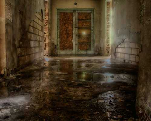 A dimly lit corridor with a wet floor, leading to a boarded-up door at the end. Walls show signs of decay.