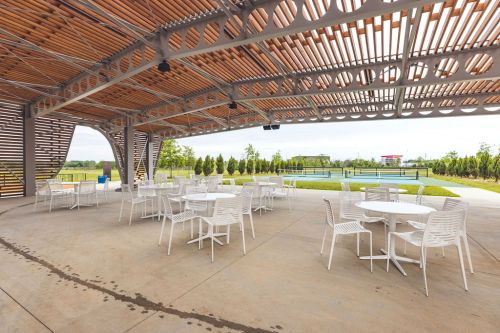 Outdoor seating area with white tables and chairs under a wooden canopy, overlooking a green landscape and water feature.