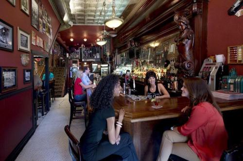A lively bar scene with patrons chatting at the counter, vintage decor, and a mirrored backdrop.