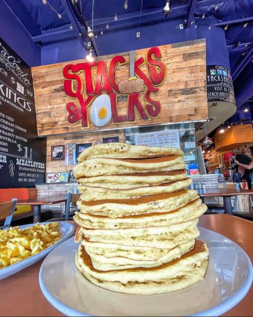 A towering stack of pancakes in front of a colorful "Stacks & Yolks" sign in a vibrant restaurant setting.