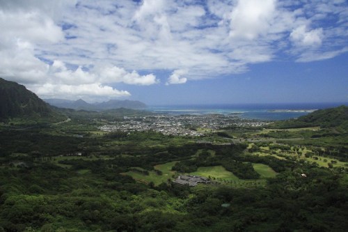 A panoramic view of a lush green valley with a coastal town and ocean in the distance under a partly cloudy sky.