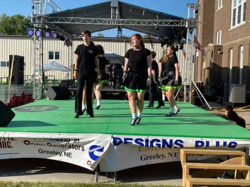 A group of performers in black and green outfits dance on a stage during an outdoor event.