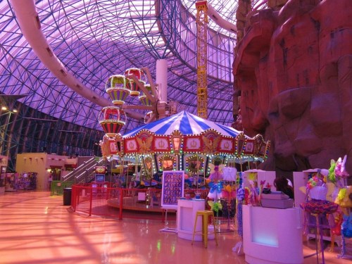 Colorful indoor amusement park featuring a carousel, rides, and vibrant decorations under a glass ceiling.