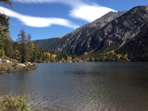 A serene lake surrounded by mountains and trees, with clear blue skies and wispy clouds above.