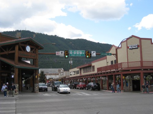 A street view of a small town with shops, traffic lights, and mountains in the background under a cloudy sky.