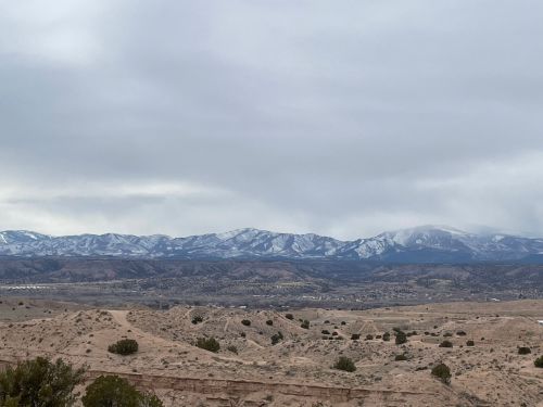 A panoramic view of snow-capped mountains under a cloudy sky, with a dry landscape in the foreground.