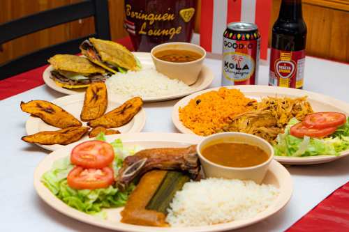 A spread of Caribbean dishes including rice, plantains, tacos, and stews, served with drinks on a table.