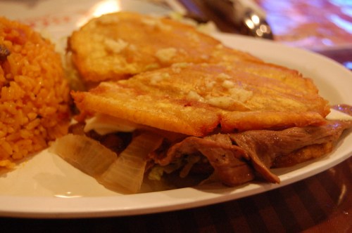 A close-up of a sandwich made with fried plantains, filled with meat, lettuce, and mayonnaise, served with rice.