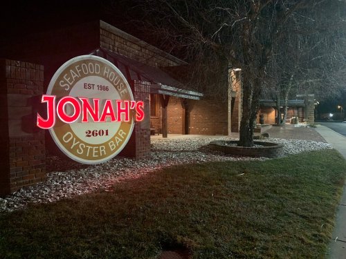 A nighttime view of Jonah's Oyster Bar sign, illuminated in red, with a brick building and landscaped area nearby.