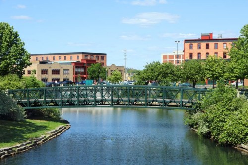 A green bridge spans a calm river, with brick buildings and trees lining the banks under a clear blue sky.