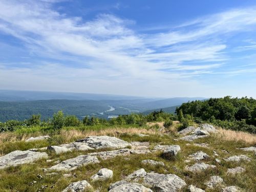 A scenic view from a rocky hilltop, overlooking a valley with a river and distant mountains under a blue sky.