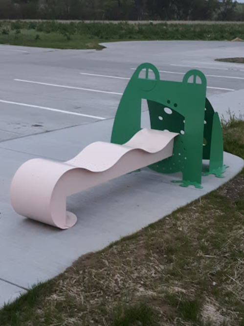 A whimsical green frog-shaped bench with a wavy pink seat in a parking lot setting.