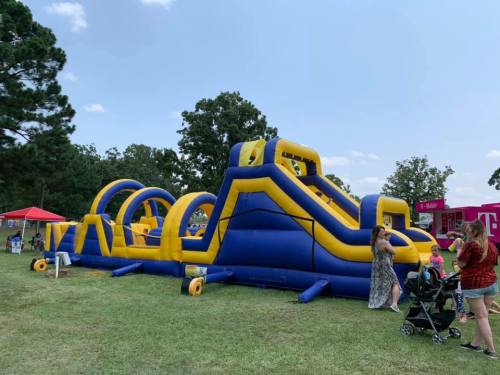 A large blue and yellow inflatable obstacle course set up on grass, with people enjoying the attraction nearby.