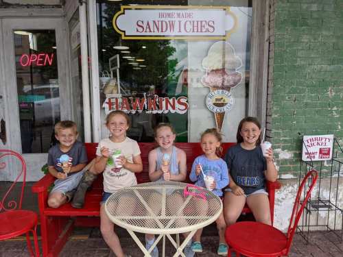 Five children sit on a red bench outside an ice cream shop, enjoying colorful ice cream cones and cups.
