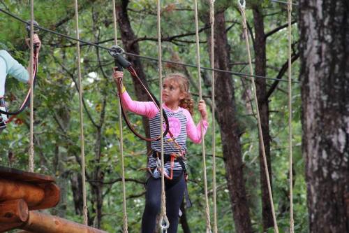 A young girl in a harness navigates a ropes course in a forested area, focused on her path ahead.