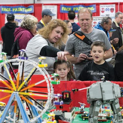 A family admires a colorful LEGO display featuring a Ferris wheel and a large robot at a crowded event.