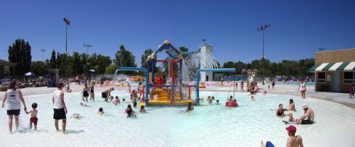 A busy water park scene with families enjoying a splash pad, water slides, and sunny weather.