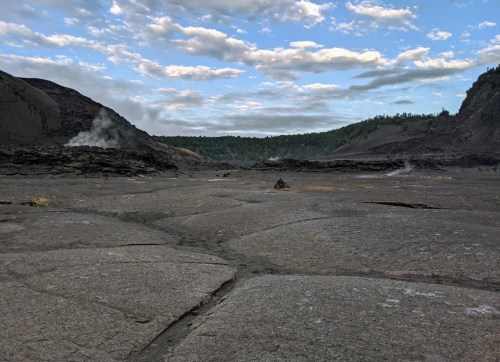 A barren volcanic landscape with steam rising from the ground, under a cloudy sky and surrounded by green hills.