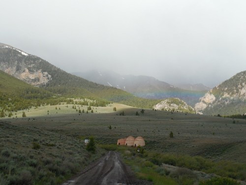 A misty landscape with rolling hills, a dirt road, and two small structures under a faint rainbow in the distance.