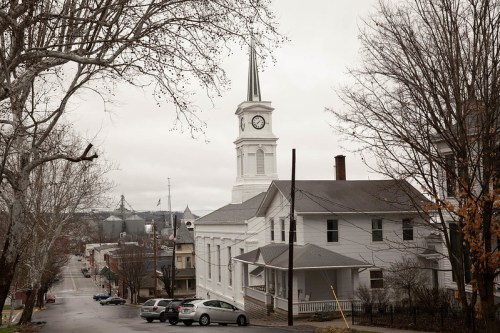 A white church with a clock tower stands on a street lined with bare trees, under a cloudy sky.
