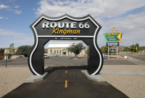 Sign for Route 66 in Kingman, Arizona, with a visitor center sign in the background and a clear blue sky.