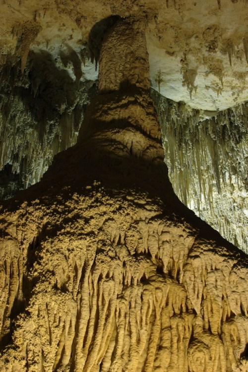 A towering stalagmite in a dimly lit cave, surrounded by intricate rock formations and stalactites above.