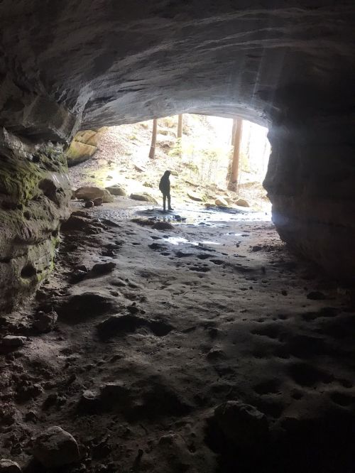 A person stands in a cave, silhouetted against a bright opening, surrounded by rocky terrain and natural light.