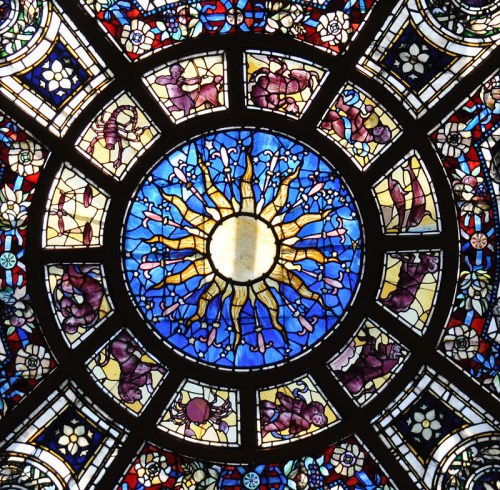 Colorful stained glass window featuring a central sun surrounded by intricate floral and figure designs.