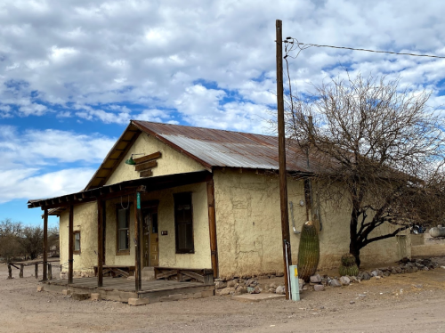 A weathered adobe house with a metal roof, surrounded by sparse vegetation and a cloudy blue sky.