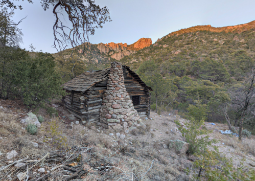 A rustic log cabin with a stone chimney, surrounded by trees and mountains at dusk.