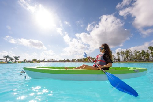 A woman in a kayak paddles on a bright blue lake under a sunny sky with fluffy clouds.