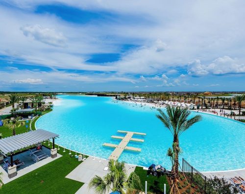 A large, bright blue lagoon-style pool surrounded by palm trees and lounge areas under a partly cloudy sky.