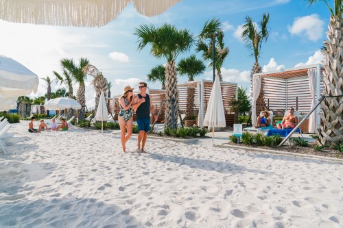 A couple walks on a sandy beach surrounded by palm trees and cabanas, with people relaxing in the background.