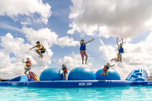 Children jumping joyfully on a large inflatable in a pool, with fluffy clouds in the blue sky above.