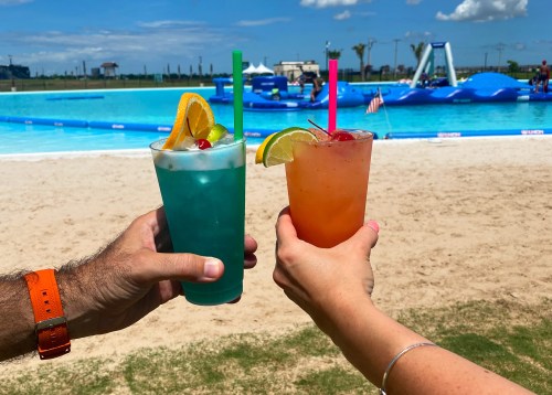 Two colorful cocktails are raised in a toast against a sunny beach backdrop with a pool and inflatable attractions.