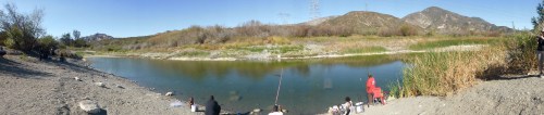 A panoramic view of a riverbank with people fishing, surrounded by greenery and mountains under a clear blue sky.